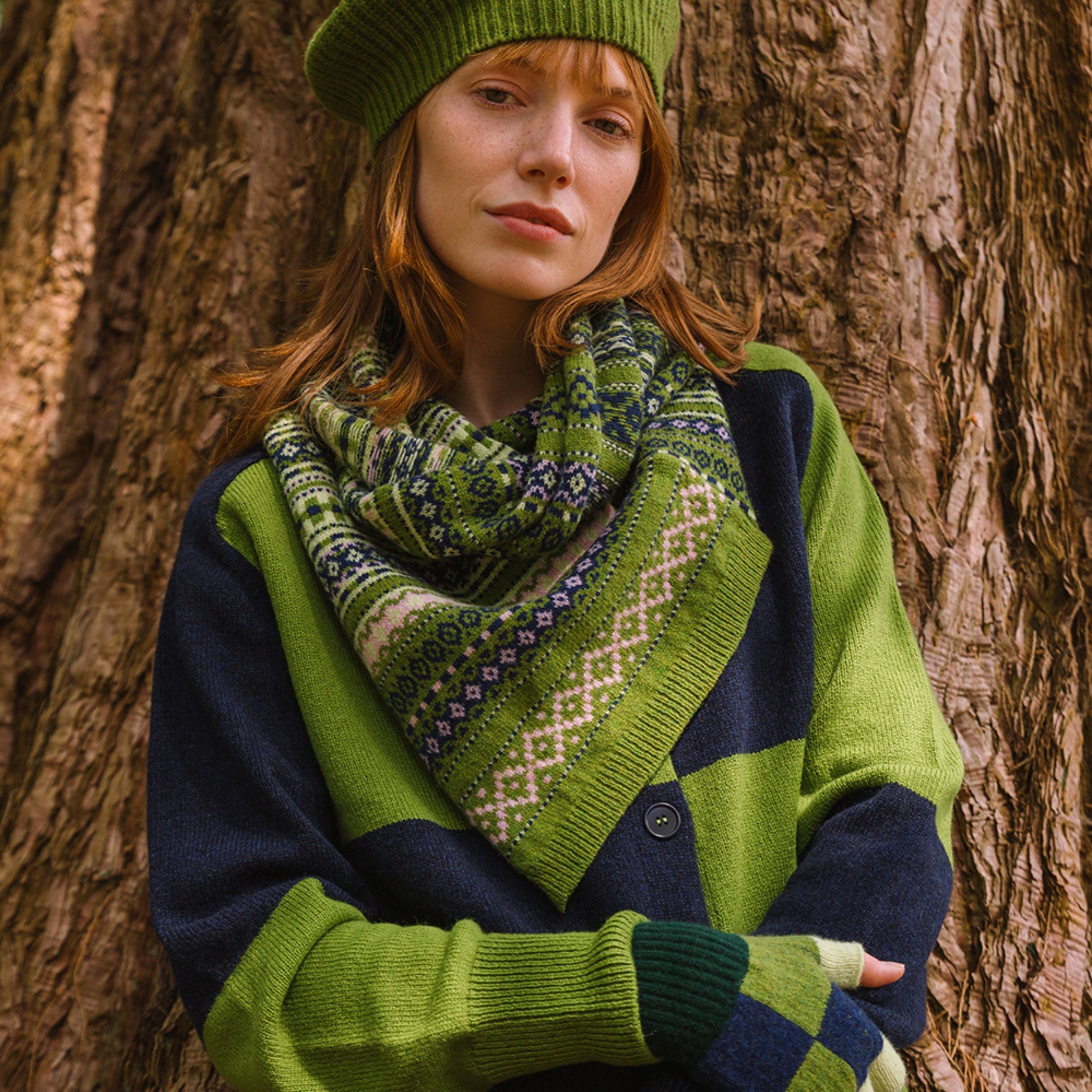 Woman wearing a green sweater and patterned scarf standing against a tree.