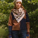 Person wearing a patterned scarf, brown and blue cardigan, and beige knit hat outdoors.