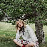 A woman crouches on grass beneath a leafy fruit tree, wearing a cream knit sweater, a red and white plaid skirt, and red socks.