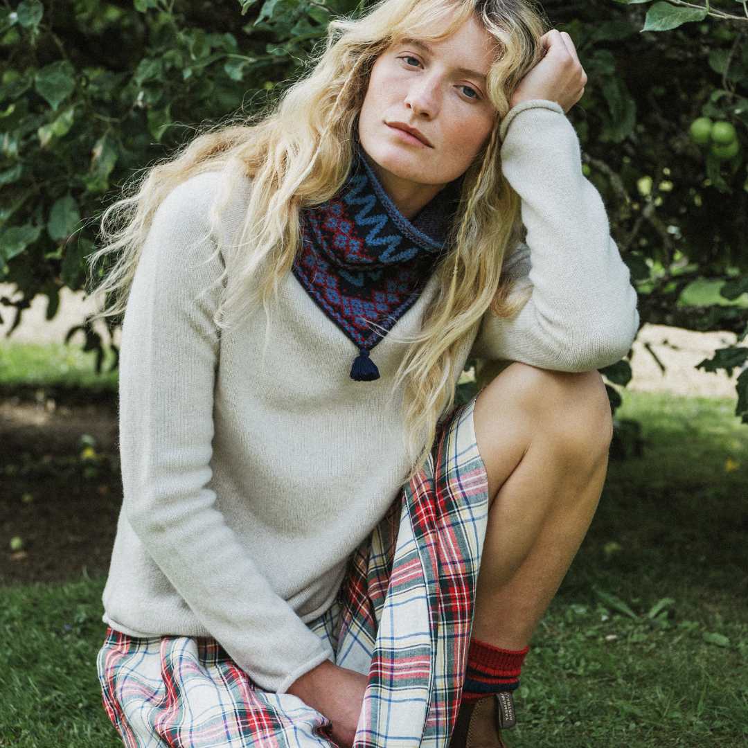 A woman crouching outdoors in front of leafy trees, wearing a cream knit sweater, a dark patterned neck snood, and a red, white, and blue plaid skirt.