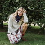 A woman crouches on grass in front of leafy trees, wearing a cream knit sweater, a patterned blue and purple neck scarf, and a red, white, and blue plaid skirt.