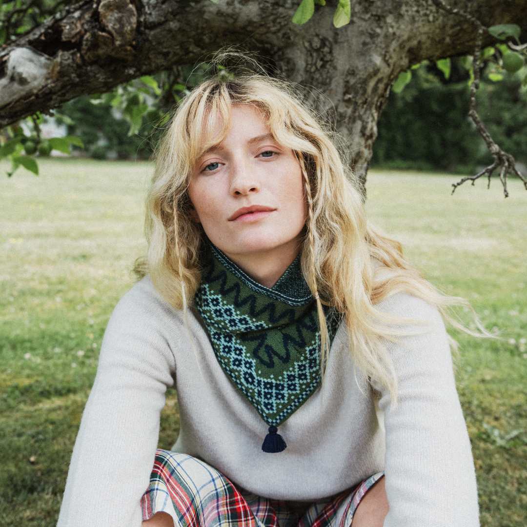 A woman with blonde hair sits outdoors under a tree wearing a light beige knit sweater, a dark green patterned bandana around her neck, and plaid pants.