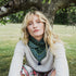 A woman with blonde hair sits outdoors under a tree wearing a light beige knit sweater, a dark green patterned bandana around her neck, and plaid pants.