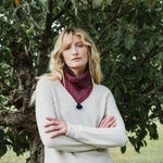A woman with blonde hair stands outdoors in front of a leafy tree, wearing a light beige knit sweater and a red patterned neck gaiter with a dark blue tassel.