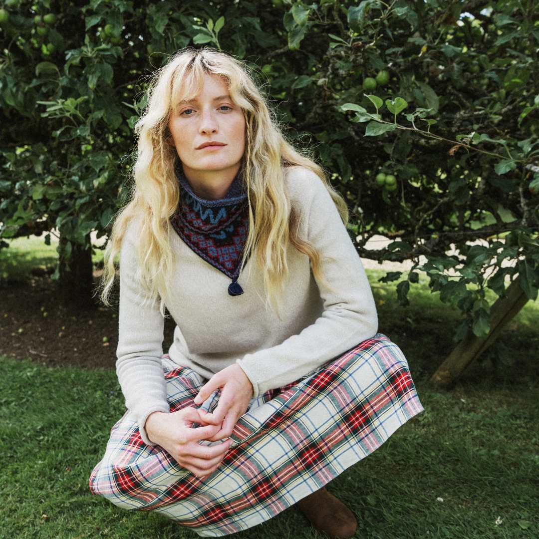 A woman with long blonde hair crouches outdoors on grass in front of leafy trees, wearing a cream knit sweater, a dark patterned neck gaiter, and a red, green, and white plaid skirt.