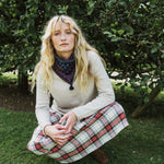 A woman with long blonde hair crouches outdoors on grass in front of leafy trees, wearing a cream knit sweater, a dark patterned neck gaiter, and a red, green, and white plaid skirt.