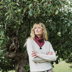 A woman with blonde hair stands in front of a leafy tree wearing a light beige knit sweater and a red patterned snood with a dark tassel. She has her arms crossed and is looking off to the side.