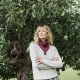 A woman with blonde hair stands in front of a leafy tree wearing a light beige knit sweater and a red patterned snood with a dark tassel. She has her arms crossed and is looking off to the side.