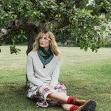 A woman sits on a grassy lawn under a leafy tree, wearing a cream knit sweater, a green patterned snood, a red and white plaid skirt, and red socks.