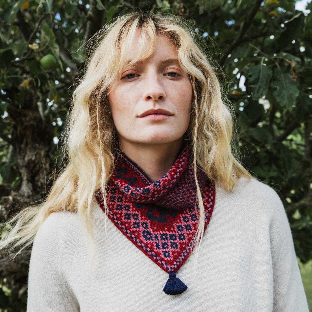A woman with blonde hair wearing a red and navy patterned knit neck warmer with a tassel, photographed outdoors in front of green foliage.