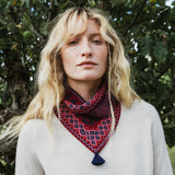 A woman with blonde hair wearing a red and navy patterned knit neck warmer with a tassel, photographed outdoors in front of green foliage.