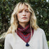 A woman with blonde hair wearing a red and navy patterned knit neck warmer with a tassel, photographed outdoors in front of green foliage.