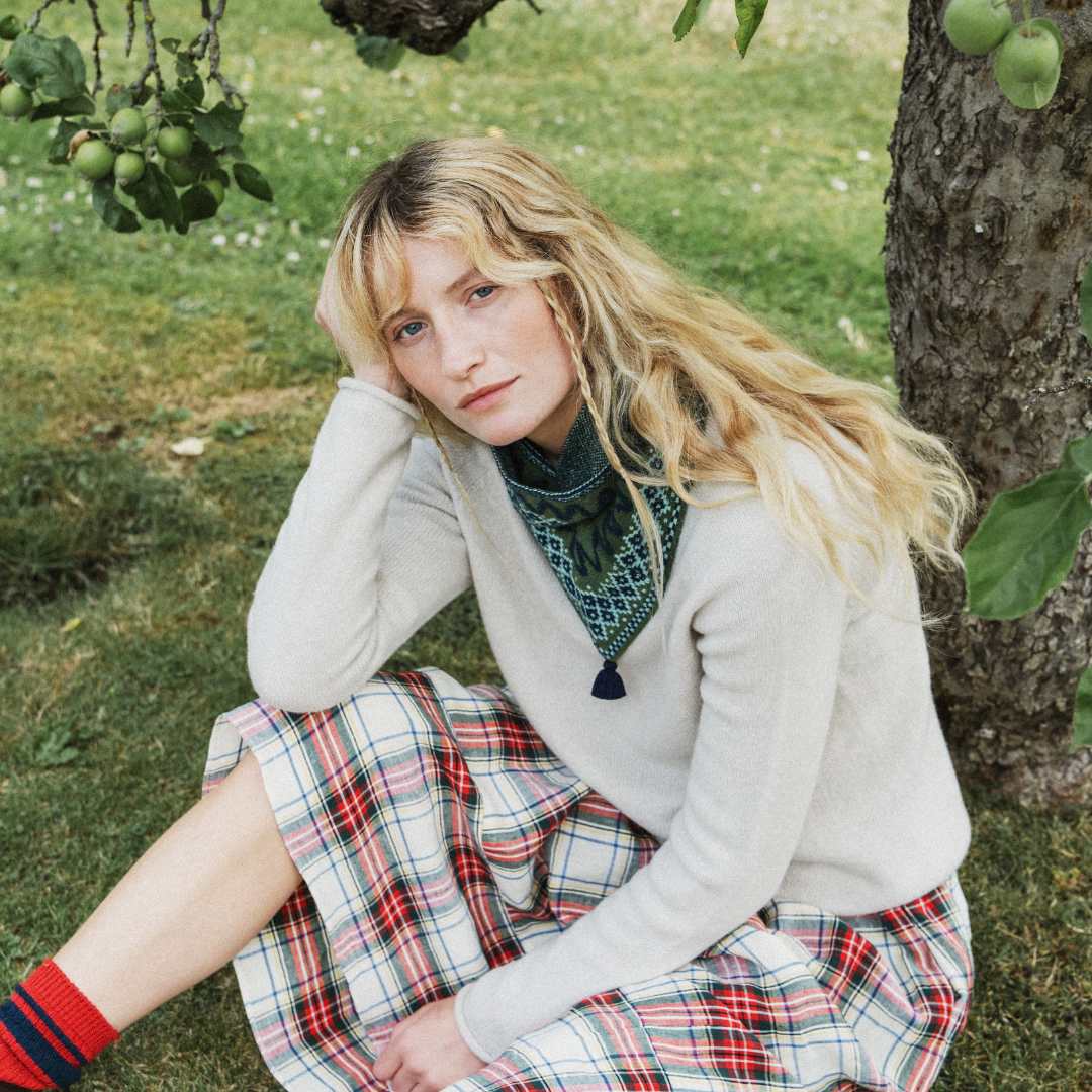 A woman sits on grass outdoors near a tree, wearing a cream knit cardigan, a patterned green neck scarf, and a red, white, and blue plaid skirt. She has long blonde hair and is wearing red striped socks.