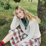 A woman sits on grass outdoors near a tree, wearing a cream knit cardigan, a patterned green neck scarf, and a red, white, and blue plaid skirt. She has long blonde hair and is wearing red striped socks.
