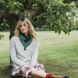 A woman sits on the grass leaning against a tree outdoors, wearing a cream knit sweater, a green patterned snood, and a red, white, and blue plaid skirt.