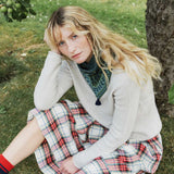 A woman sits on grass outdoors near a tree, wearing a light beige cardigan over a green patterned top and a red, white, and blue plaid skirt. She has a green knit scarf and red socks.