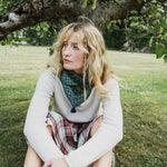 A woman sitting outdoors under a tree on a grassy lawn, wearing a light gray knit sweater, a green and navy patterned bandana around her neck, and a red plaid skirt.