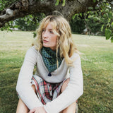 A woman sitting outdoors under a tree on a grassy lawn, wearing a light gray knit sweater, a green and navy patterned bandana around her neck, and a red plaid skirt.