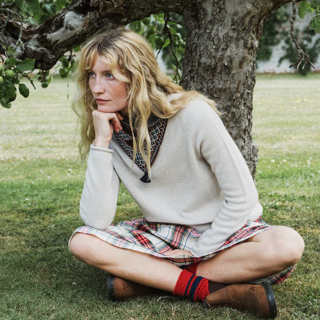 A woman sits cross-legged on grass in front of a tree, wearing a cream knit sweater, a plaid skirt, red socks, a green snood and brown shoes.
