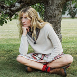 A woman sits cross-legged on grass in front of a tree, wearing a cream knit sweater, a plaid skirt, red socks, a green snood and brown shoes.