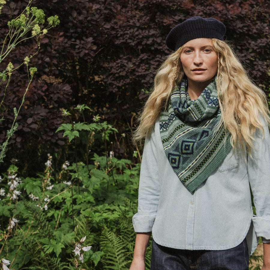 A woman standing outdoors in a garden wearing a dark navy beret, a light sage green button-up shirt, dark jeans, and a green patterned knit scarf.