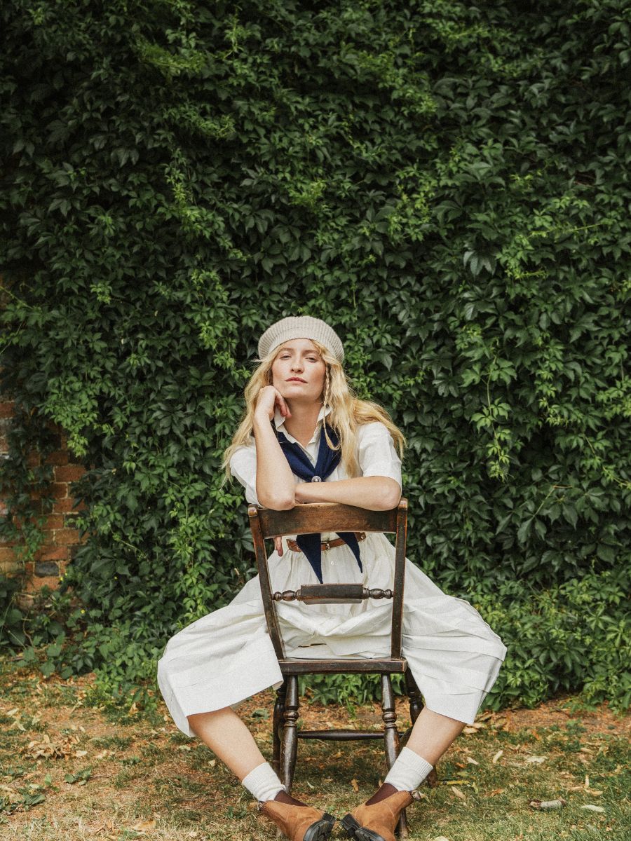 A woman sits on a wooden chair outdoors in front of a dense green ivy-covered wall. She is wearing a white dress, a beige beret, and a navy blue neck scarf.