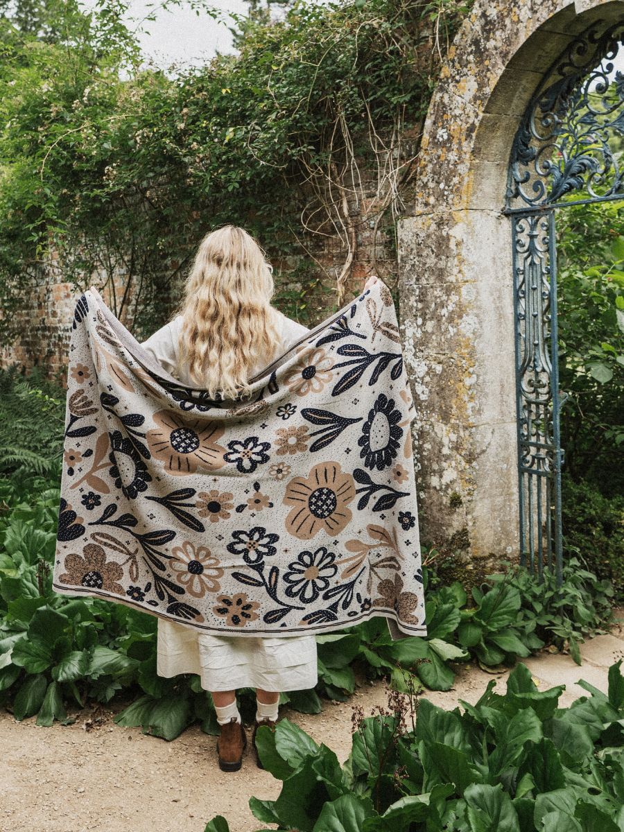 A woman stands with her back to the camera in a garden courtyard, holding up a large patterned blanket or throw. The blanket features a cream background with a floral and botanical design in black, brown, and muted blue.