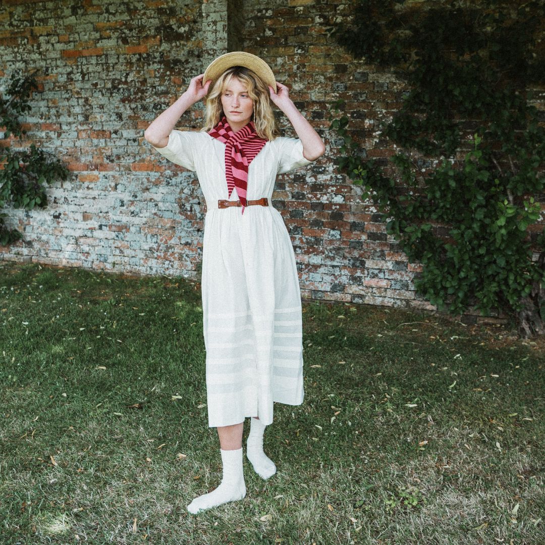 A woman stands outdoors on grass in front of a brick wall, wearing a cream midi dress with a red and white striped neck scarf, a brown belt, white socks, and a straw hat.