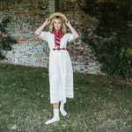 A woman stands outdoors on grass in front of a brick wall, wearing a cream midi dress with a red and white striped neck scarf, a brown belt, white socks, and a straw hat.