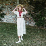 A woman stands outdoors on grass in front of a brick wall, wearing a cream midi dress with a red and white striped neck scarf, a brown belt, white socks, and a straw hat.