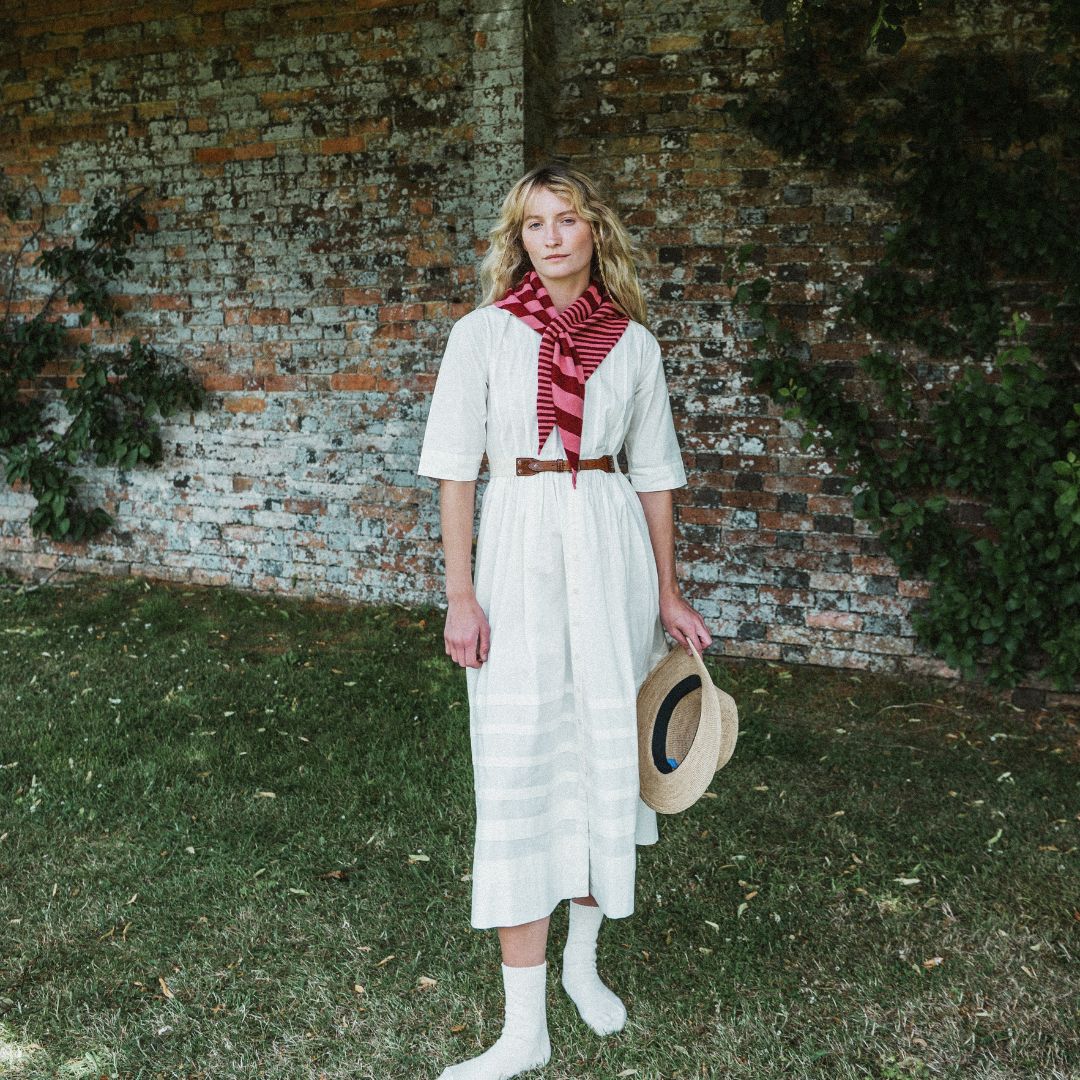 A woman stands outdoors in front of a weathered brick wall and grassy ground, wearing a cream-colored midi dress with a red and white striped neck scarf, a thin brown belt, white socks, and holding a straw hat.