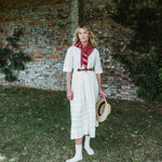 A woman stands outdoors in front of a weathered brick wall and grassy ground, wearing a cream-colored midi dress with a red and white striped neck scarf, a thin brown belt, white socks, and holding a straw hat.