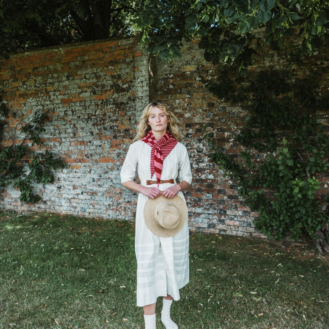 A woman stands outdoors in front of a weathered brick wall and greenery, wearing a cream-colored long dress with a red and white striped neck scarf and holding a straw hat.