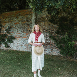 A woman stands outdoors in front of a weathered brick wall and greenery, wearing a cream-colored long dress with a red and white striped neck scarf and holding a straw hat.