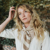 A woman with long wavy blonde hair wearing a cream-colored shirt and a beige and white striped knit scarf, posing against a rustic brick wall with green foliage.