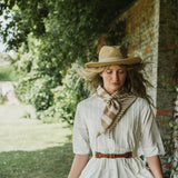 A woman outdoors wearing a cream short-sleeve dress with a brown belt, a beige straw hat, and a beige-and-white striped scarf, standing in front of a rustic brick wall and greenery.