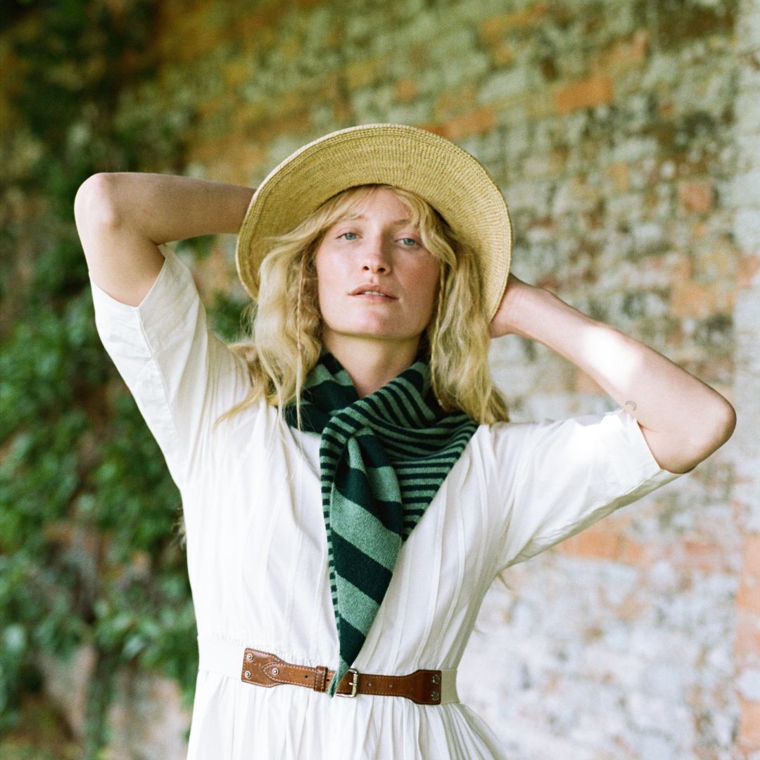 A woman wearing a straw hat, a white dress, and a green striped scarf poses with her hands behind her head against a rustic stone wall background.