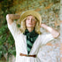 A woman wearing a straw hat, a white dress, and a green striped scarf poses with her hands behind her head against a rustic stone wall background.