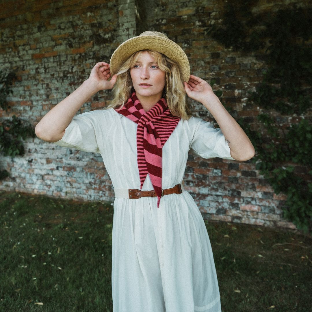 A woman wearing a white dress, brown belt, straw hat, and red striped scarf stands in front of a rustic brick wall outdoors.