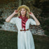 A woman wearing a white dress, brown belt, straw hat, and red striped scarf stands in front of a rustic brick wall outdoors.
