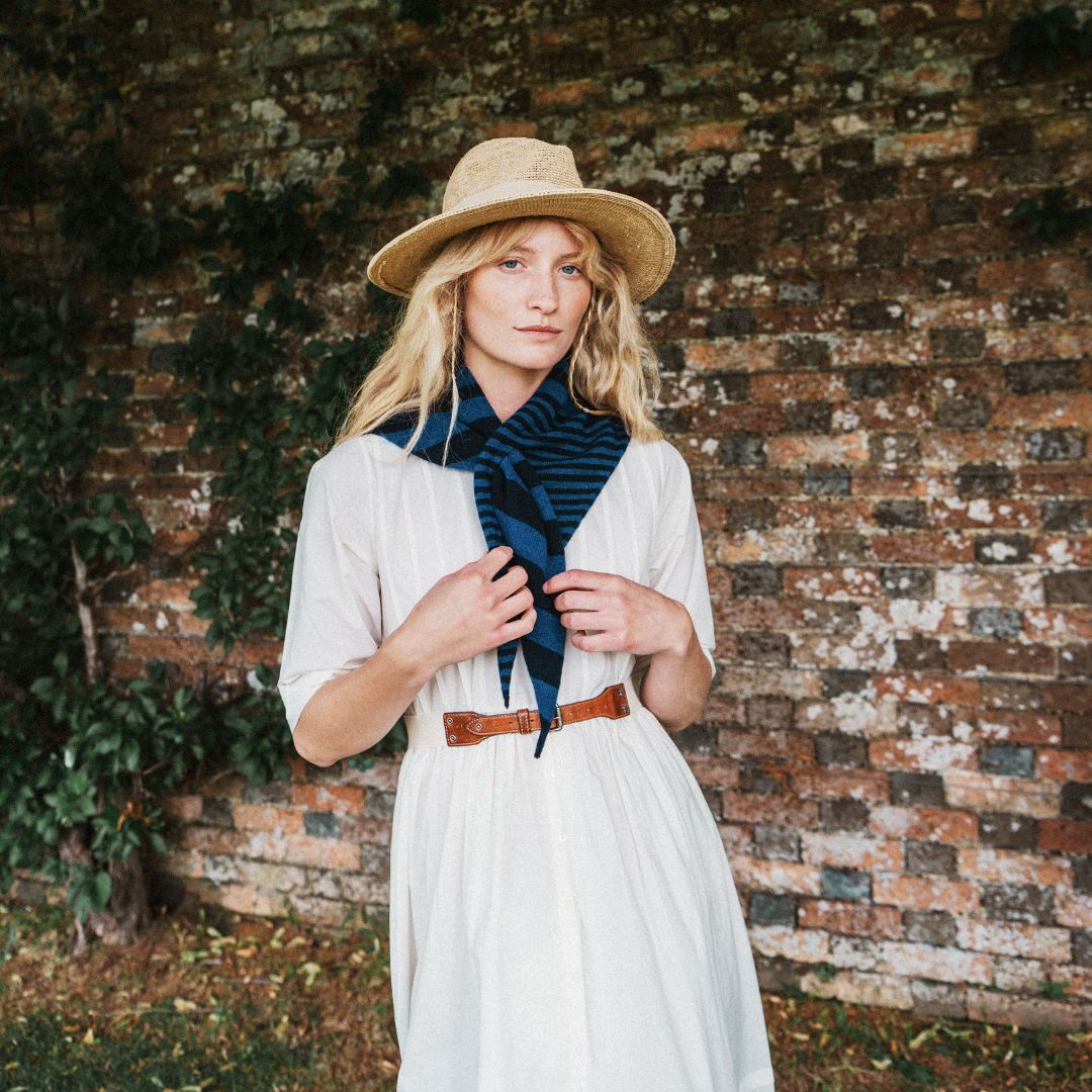 A woman stands outdoors in front of a weathered brick wall wearing a cream-colored dress, a wide-brimmed straw hat, and a dark blue striped scarf tied around her neck.