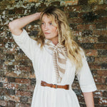 A woman with long blonde hair stands against a weathered brick wall, wearing a white dress with a brown belt and a beige and white striped scarf tied around her neck.