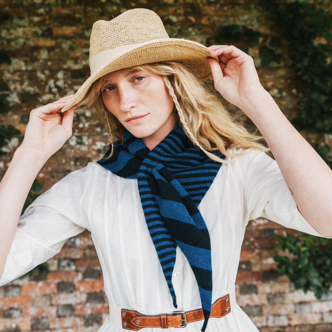 A woman wearing a straw cowboy hat, a white dress, and a navy blue and black striped scarf, posing in front of a rustic brick wall.