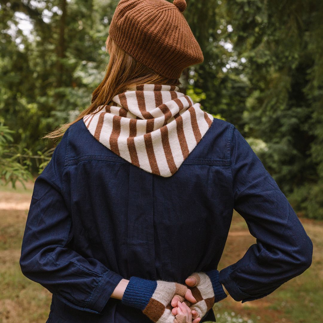 Person wearing a blue jacket, brown beanie, and striped scarf standing in a forest.