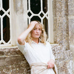 A woman with blonde hair leans against a stone wall in front of a decorative window, wearing a white collared shirt and a light beige wrap tied around her waist.