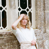A woman with blonde hair leans against a stone wall in front of a decorative window, wearing a white collared shirt and a light beige wrap tied around her waist.