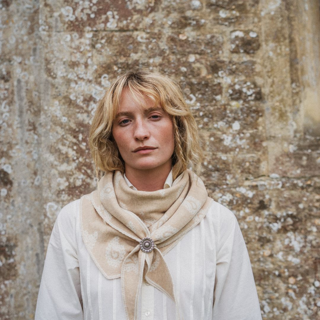 A woman with blonde hair wearing a cream-colored blouse and a beige patterned scarf with a decorative brooch, standing against a textured stone wall.