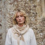 A woman with blonde hair wearing a cream-colored blouse and a beige patterned scarf with a decorative brooch, standing against a textured stone wall.
