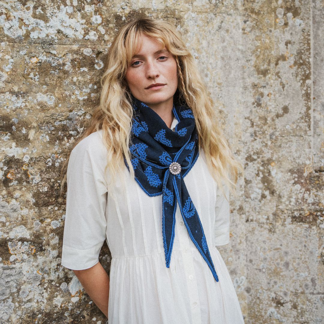 A woman with long blonde hair stands against a textured stone wall wearing a white dress and a navy blue patterned scarf tied around her neck with a scarf slider..