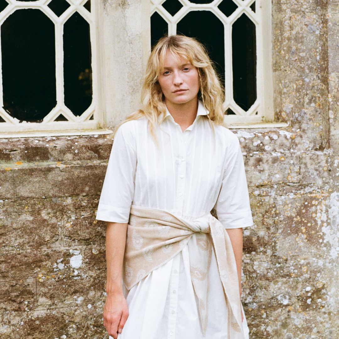 A woman with blonde hair stands in front of a stone wall and window, wearing a white short-sleeve button-up shirt dress with a beige sash tied at the waist.